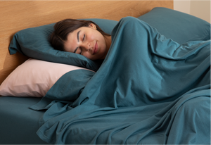 Woman sleeping peacefully in teal It-Fits Australian cotton jersey sheets, wrapped in a doona, with soft pink and teal pillows on a wooden bed.