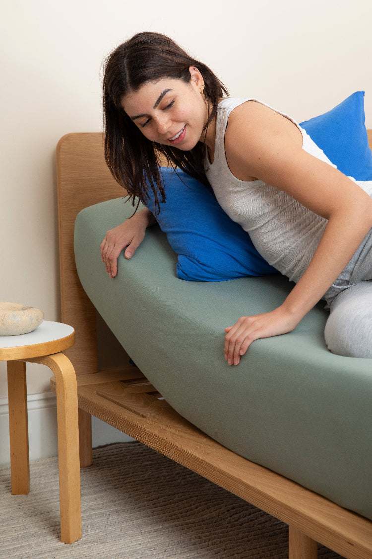 A woman smiling contentedly on an adjustable bed, with a sage green fitted sheet perfectly snug and a royal blue pillowcase resting on the pillow. The bed features a wooden frame with a matching wooden side table beside it.